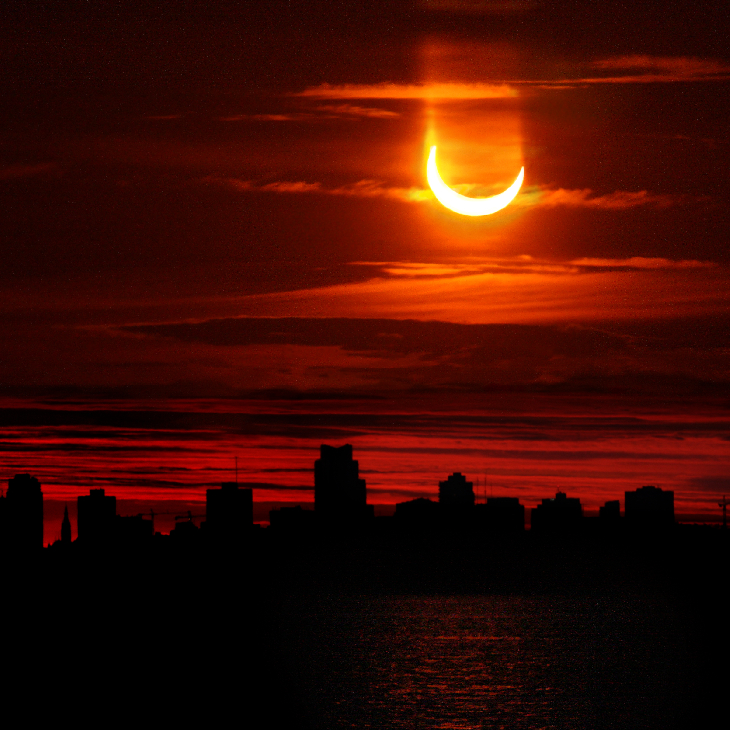 Crescent eclipsed sun setting over a city skyline in a fiery red sky
